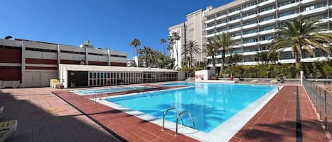 A swimming pool with a tiled deck, a parking garage, and a multi-story building with balconies.