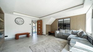 Un salon moderne avec un canapé gris, un banc en bois, une grande horloge et une vue sur l’extérieur.