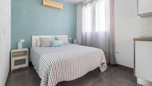Een slaapkamer met een bed, nachtkastje, raam met gordijnen met patroon, en een airconditioning.
