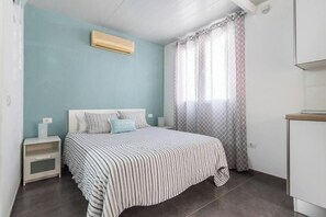 Ein Schlafzimmer mit Bett, Nachttisch, Fenster mit gemusterten Vorhängen und einer Klimaanlage.