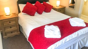 A neatly made bed with red pillows, white linens, and a red throw blanket. There are two bedside tables with lamps, a wooden nightstand with drawers, and a mirror on the wall. - Brewery Lodge Sleeps 4 (Lydney)