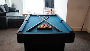 Una mesa de billar en una sala de estar con un sofá y un televisor al fondo.