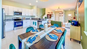 A modern kitchen with white cabinets, a microwave, and an oven. There is a dining area with a wooden table set for four, blue chairs, and a chandelier. - Designer Home with Community Pool Near Apollo Beach (Ruskin)