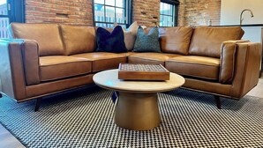 A brown leather sectional sofa with a round coffee table in a room with brick walls and large windows. - Brick & Bed - Two Bedroom Riverfront Loft (St. Louis)