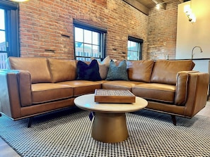 A brown leather sectional sofa with a round coffee table in a room with brick walls and large windows. - Brick & Bed - Two Bedroom Riverfront Loft (St. Louis)