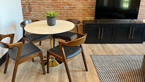 A room with a brick wall, a round table with chairs, a black cabinet, and a TV. - Brick & Bed - Two Bedroom Riverfront Loft (St. Louis)