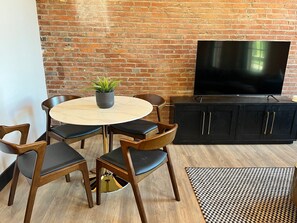 A room with a brick wall, a round table with chairs, a black cabinet, and a TV. - Brick & Bed - Two Bedroom Riverfront Loft (St. Louis)