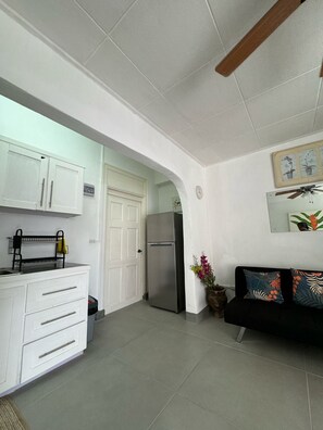 A compact kitchen with white cabinets, a stainless steel refrigerator, and a black sofa with floral cushions. - Cozy 1-bedroom 2 bathroom apartment in heart of Barbados south coast. (Bridgetown)