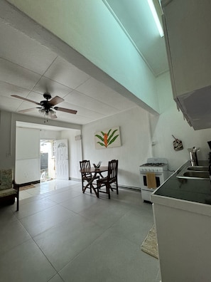 A spacious living area with a dining table, chairs, a ceiling fan, a kitchen with a stove, and a door leading outside. - Cozy 1-bedroom 2 bathroom apartment in heart of Barbados south coast. (Bridgetown)
