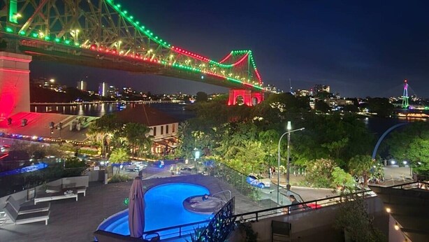 Une vue nocturne d’un pont illuminé de lumières colorées, d’un espace piscine avec des chaises longues, et d’un paysage urbain en arrière-plan.
