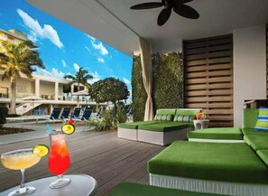 Uma área de lounge moderna ao ar livre, com assentos acolchoados verdes, um bar à beira da piscina com bebidas e vista para o edifício do hotel.