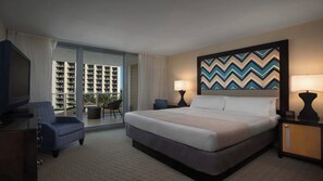 Quarto de hotel com uma cama, duas mesas de cabeceira com abajures, uma cadeira, uma televisão e uma janela grande com vista para um edifício.
