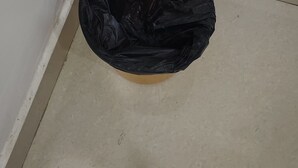 Eine schwarze Müllsäcke steht auf einem beigen Boden in einer Ecke eines Raumes mit weißen Wänden.