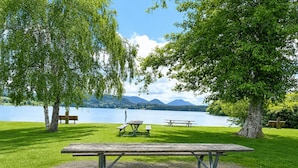 A picnic area with a table and benches, surrounded by trees and a lake with mountains in the background.