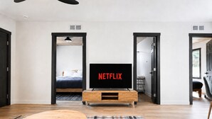 A modern living room with a wooden coffee table, a flat-screen TV displaying 'NETFLIX,' and a bed visible through an open doorway.