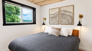 A bedroom with a bed, a window with a view of greenery, two wall-mounted lamps, and two framed pictures.
