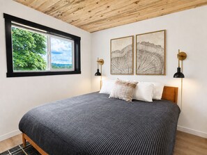 A bedroom with a bed, a window with a view of greenery, two wall-mounted lamps, and two framed pictures.