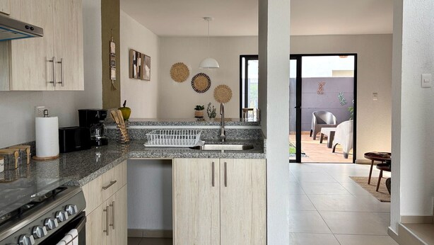 A modern kitchen with granite countertops, a built-in oven, and a dining area with wicker furniture. - Casa Bugambilia at Senderos de Opico2 | WiFi, Pool, Gym (San Juan Opico)