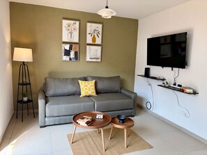 Ein Wohnzimmer mit einem grauen Sofa, zwei Beistelltischen, einer Stehlampe und einem an der Wand befestigten Fernseher.