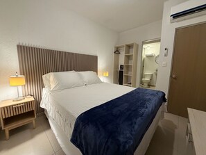 Ein Hotelzimmer mit Bett, Nachttisch, Lampe, Kleiderschrank und einem durch eine geöffnete Tür einsehbaren Badezimmer.
