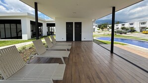 A covered outdoor seating area with wooden flooring and a view of a swimming pool and buildings in the background. - Casa Bugambilia at Senderos de Opico2 | WiFi, Pool, Gym (San Juan Opico)