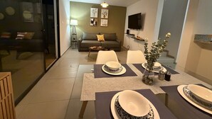 A modern living room with a sofa, a dining table set with plates, a TV mounted on the wall, and a floor lamp. - Casa Bugambilia at Senderos de Opico2 | WiFi, Pool, Gym (San Juan Opico)
