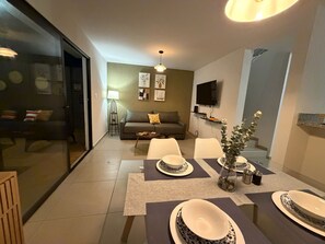 Un salon moderne avec un canapé, une table à manger dressée avec de l’assiette, un téléviseur fixé au mur et un lampadaire.