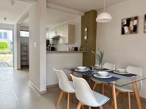 Een moderne eethoek met een glazen tafel, witte stoelen en een keuken op de achtergrond.