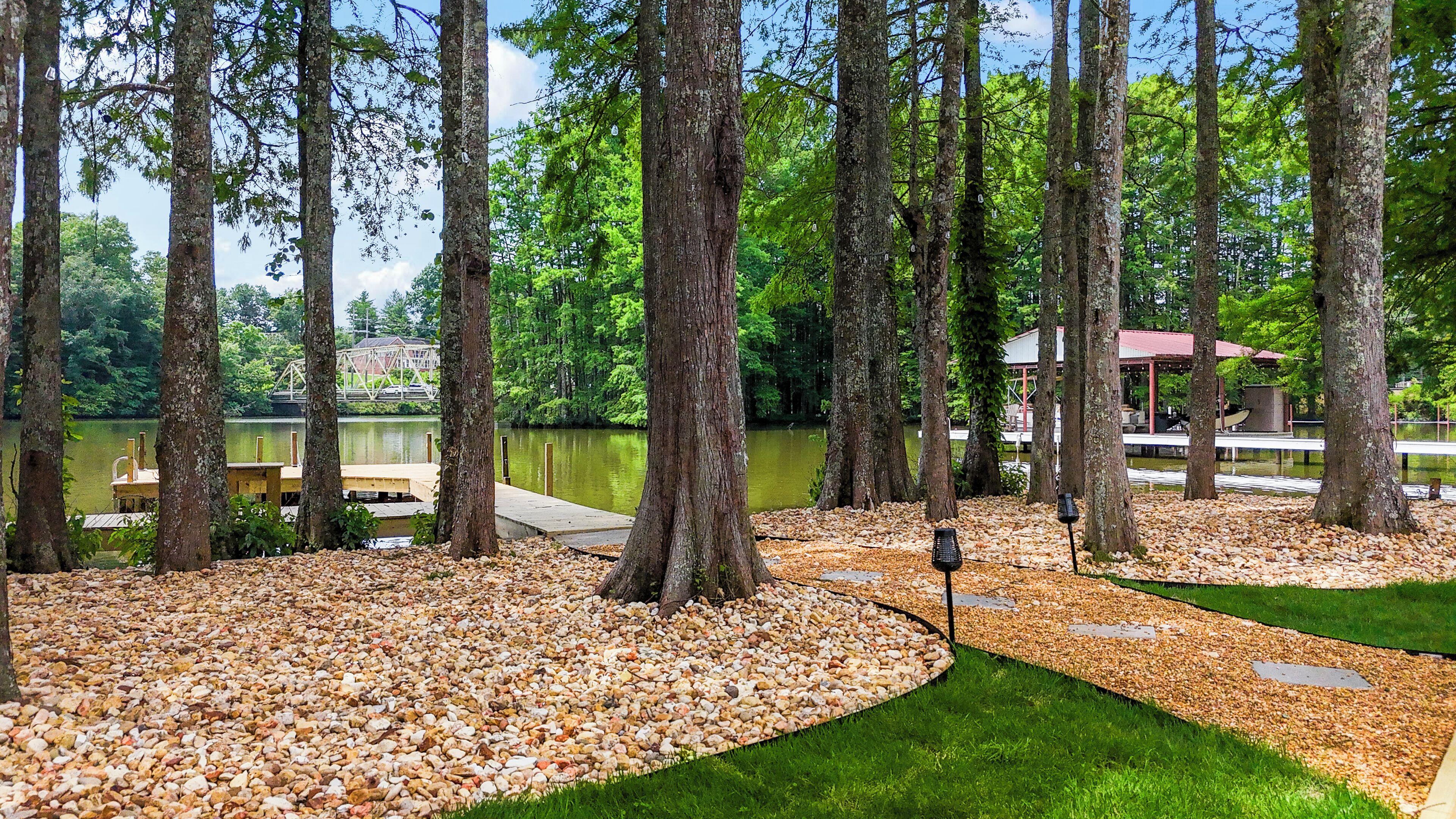 A serene lakeside setting with a wooden dock, surrounded by tall trees and pebbled paths.