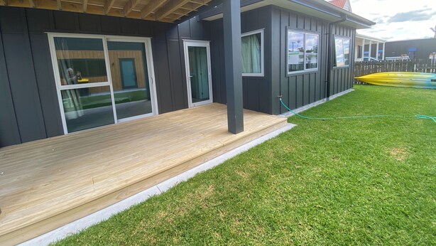 Une maison moderne à deux étages, dotée d’une terrasse en bois, de grandes portes-fenêtres coulissantes et d’une pelouse bien entretenue.