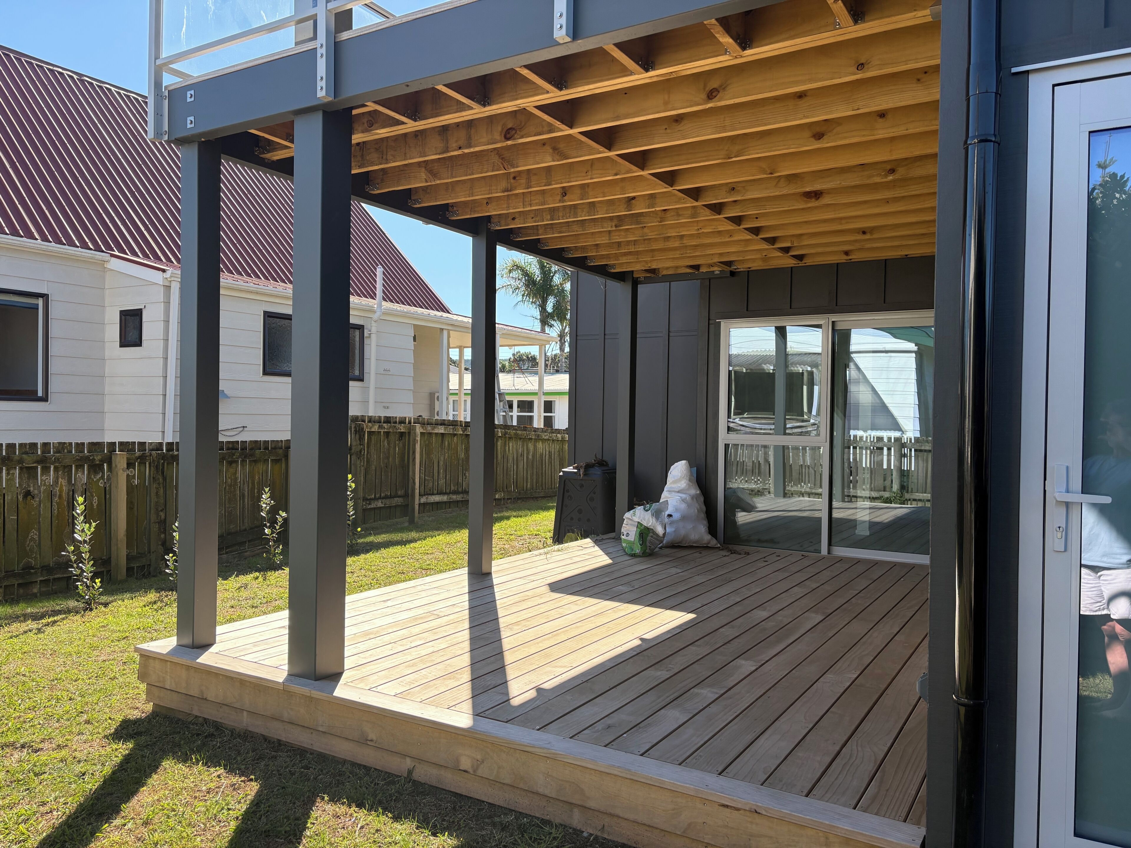 Une terrasse extérieure couverte, dotée d’un plancher en bois, de poutres métalliques et d’une porte vitrée donnant sur un jardin.