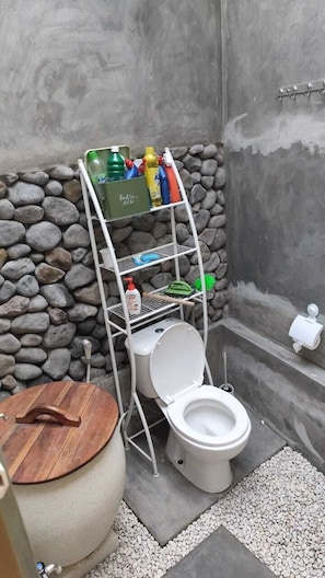 A compact bathroom with a stone wall, a white toilet, a wooden bucket, and a metal shelf with toiletries. - nDalem Riyartan Villa Kaliurang (Sleman)