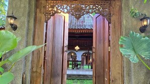 A traditional wooden door with a decorative metal frame, leading to an outdoor area with a thatched roof and stone flooring. - nDalem Riyartan Villa Kaliurang (Sleman)