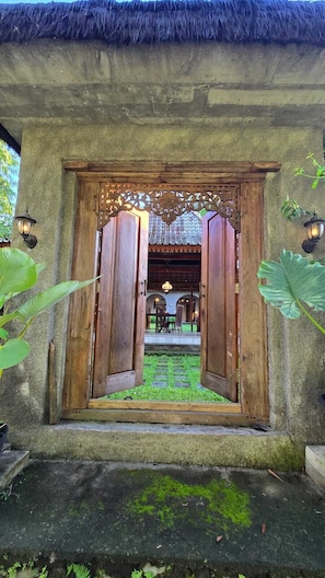 A traditional wooden door with a decorative metal frame, leading to an outdoor area with a thatched roof and stone flooring. - nDalem Riyartan Villa Kaliurang (Sleman)