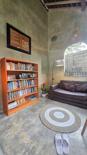A living room with a bookshelf, a sofa, a rug, and a pair of slippers. - nDalem Riyartan Villa Kaliurang (Sleman)