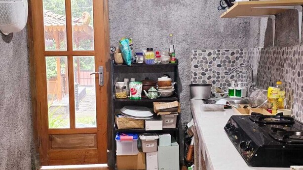 A compact kitchen with a wooden door, a shelf with various kitchen items, a countertop with a stove, and a tiled backsplash. - nDalem Riyartan Villa Kaliurang (Sleman)