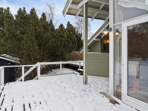 Une terrasse enneigée avec une rambarde blanche, un bâtiment en bois doté d’une porte en verre, et une forêt en arrière-plan.