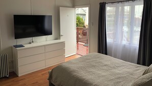 A bedroom with a bed, a TV on a dresser, a sliding door to a balcony, and a window with curtains.