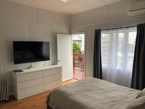 Un dormitorio con cama, televisor sobre una cómoda, puerta corrediza a un balcón y ventana con cortinas.