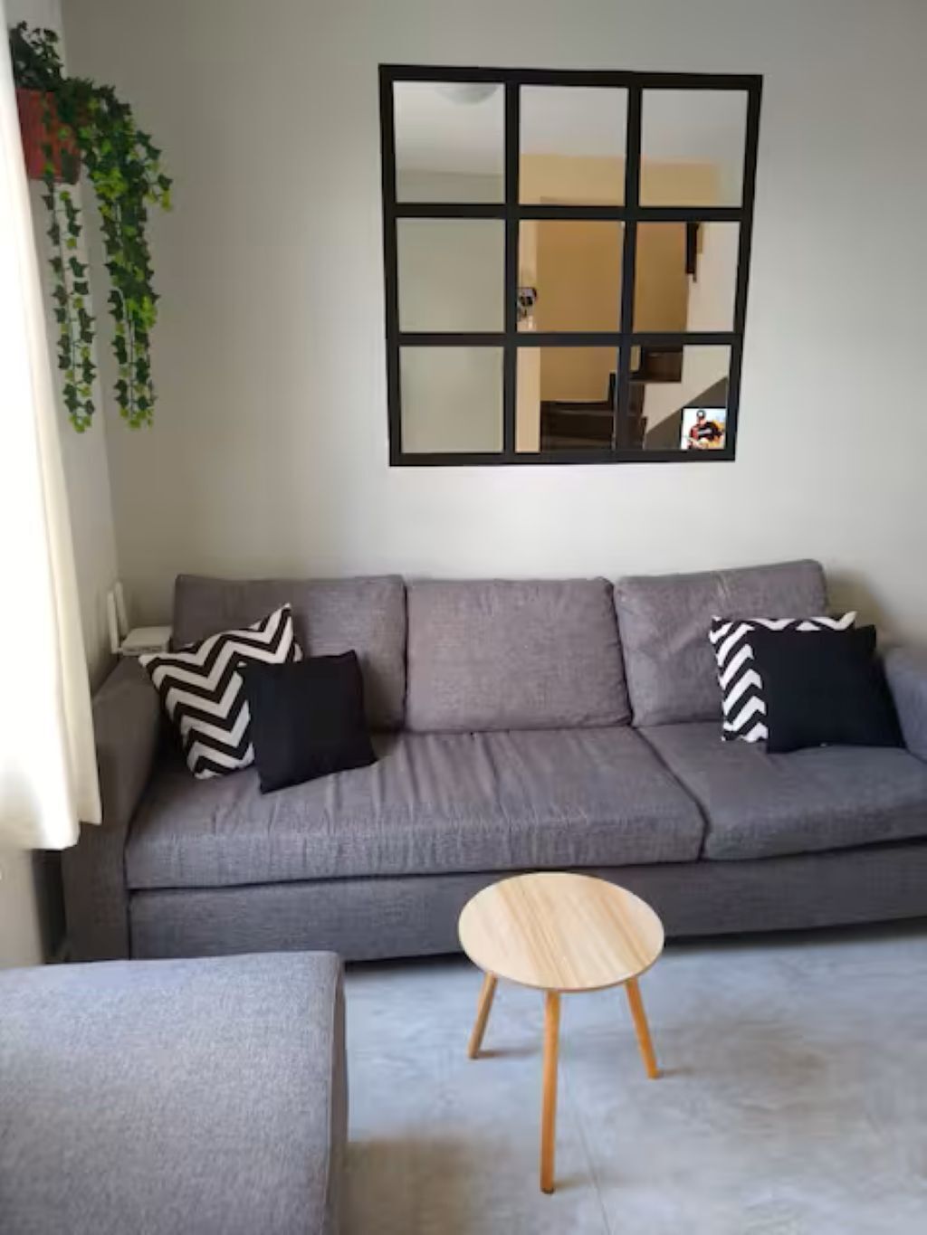 A grey sofa with black and white patterned pillows, a round wooden side table, and a mirror with a grid design on the wall.