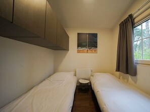 Quarto pequeno com duas camas de solteiro, um radiador, uma mesa e uma janela com cortinas.