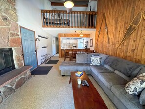 Ein gemütliches Wohnzimmer mit Kamin aus Stein, Holzbalkendecke, grauem Sofa und einem Essbereich im Hintergrund.