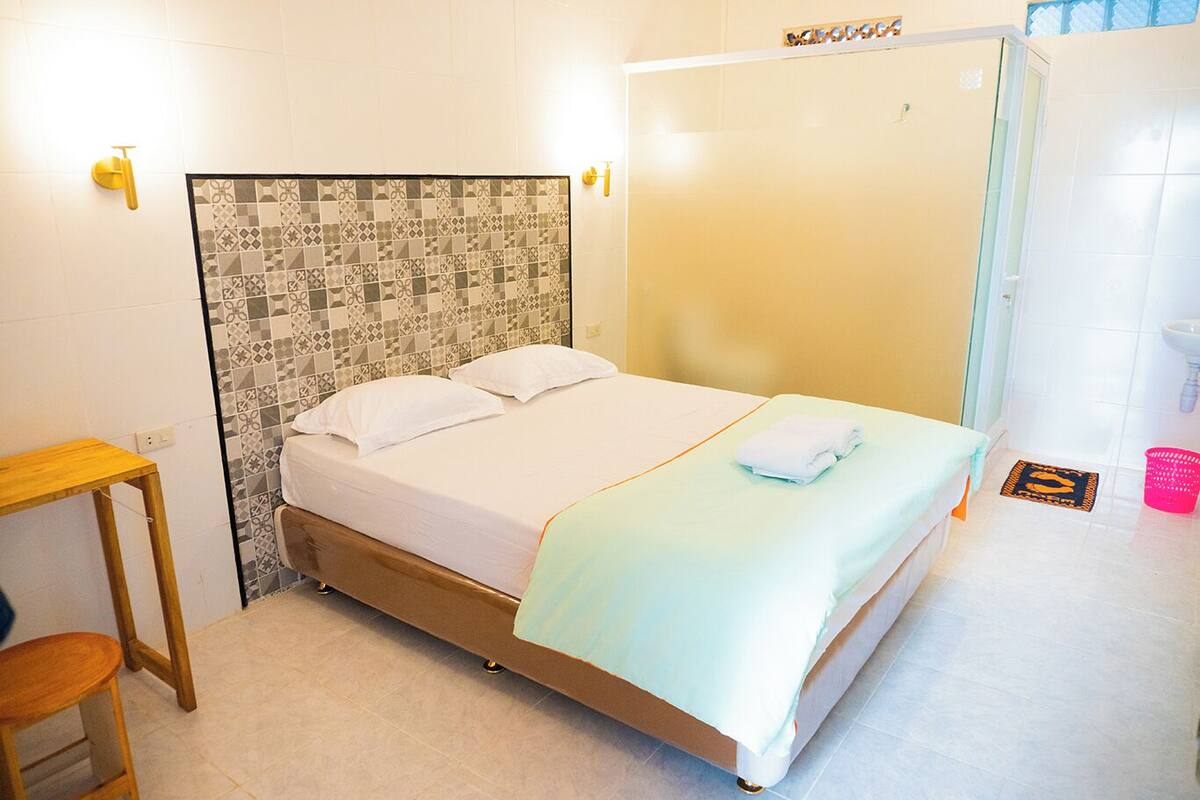 Una cama individual con cabecera estampada, una mesita de noche de madera, un baño con lavamanos blanco y zona de ducha.