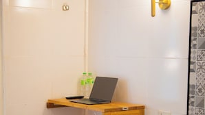 Quarto de hotel com uma cama, uma pequena mesa de madeira com um laptop, uma garrafa de água e um candeeiro fixado na parede.