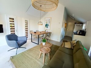 Ein modernes Wohnzimmer mit einer Couch, einem blauen Sessel, einem Esstisch mit Stühlen, einer Hängelampe und einer Küchenzeile im Hintergrund.
