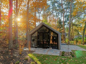 Uma casa moderna de vidro com telhado triangular, rodeada por uma floresta com folhagem de outono.