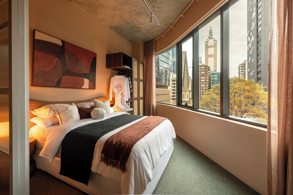 A hotel room with a large bed, a city view, a painting on the wall, and a window with curtains.
