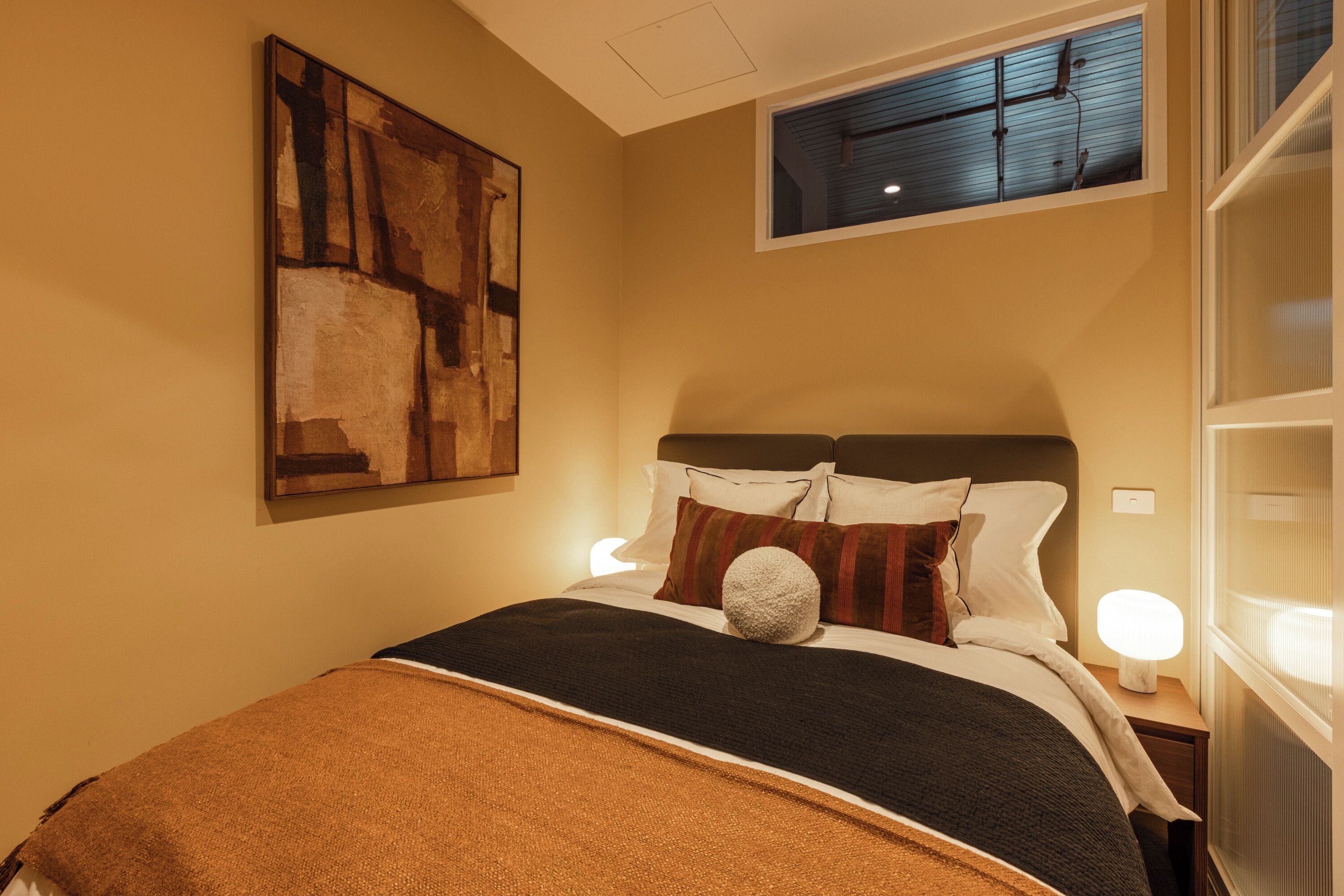 A hotel room with a bed, a painting on the wall, a window, and a nightstand with a lamp.