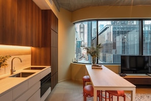 A modern kitchen with a built-in oven, a sink, and wooden cabinets. There is a dining area with a table and chairs, and a large window offering a view of the city. - The Lane House (Melbourne)