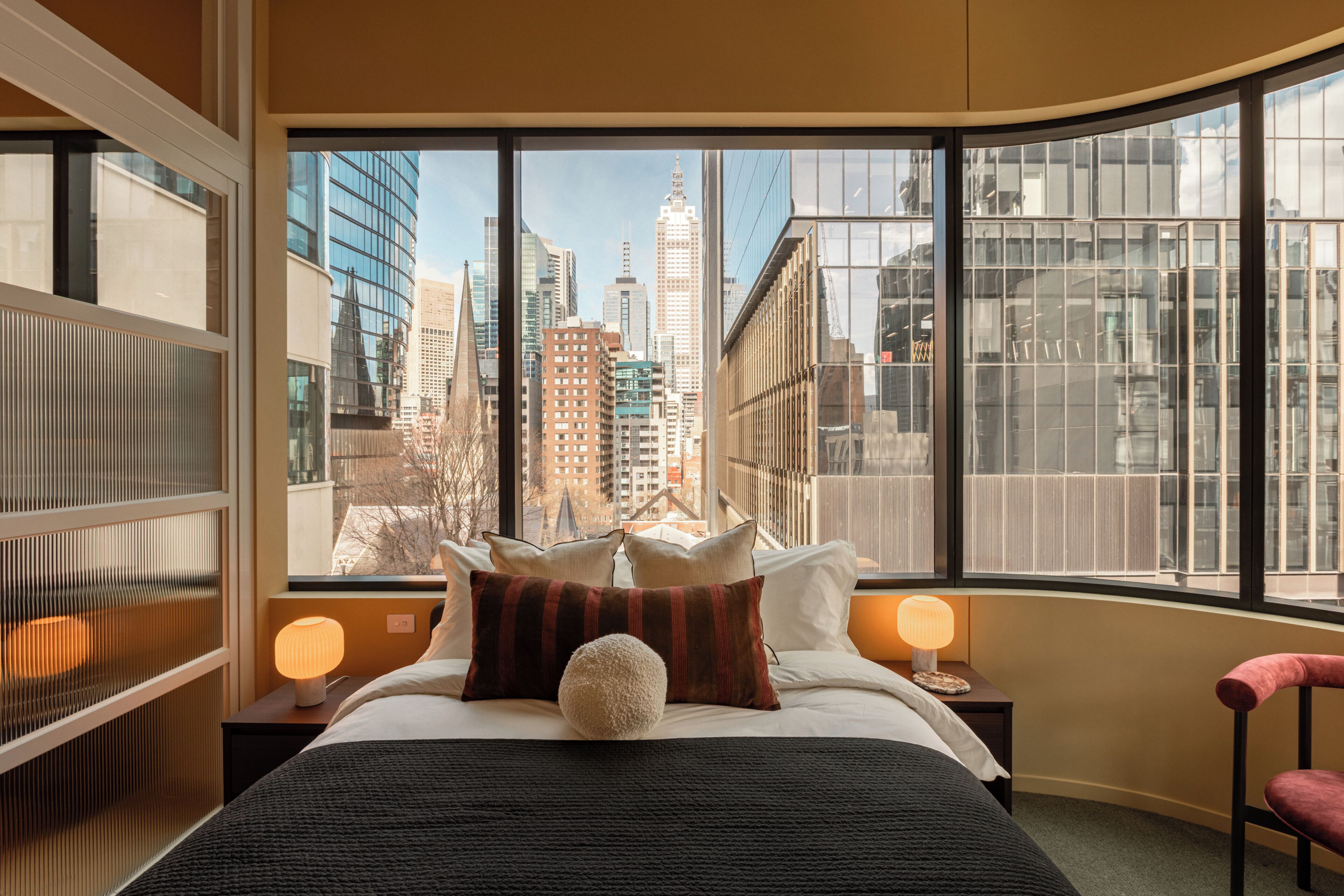 A hotel room with a large window offering a city view, a bed with pillows, a bedside lamp, and a chair.